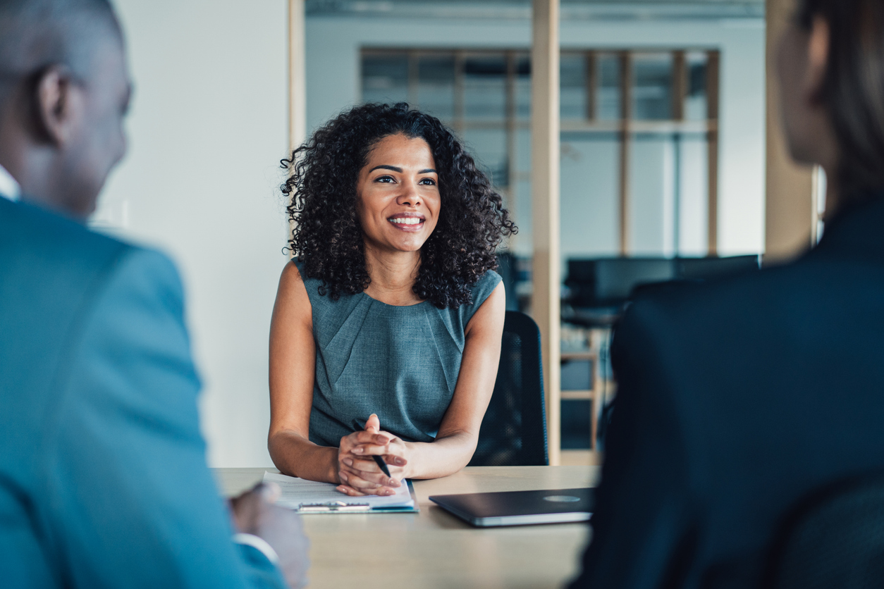 Woman sitting at conference table smiling. She has a clipboard with papers in front of her and is holding a pen. Two people are sitting across from her and are facing away from the camera