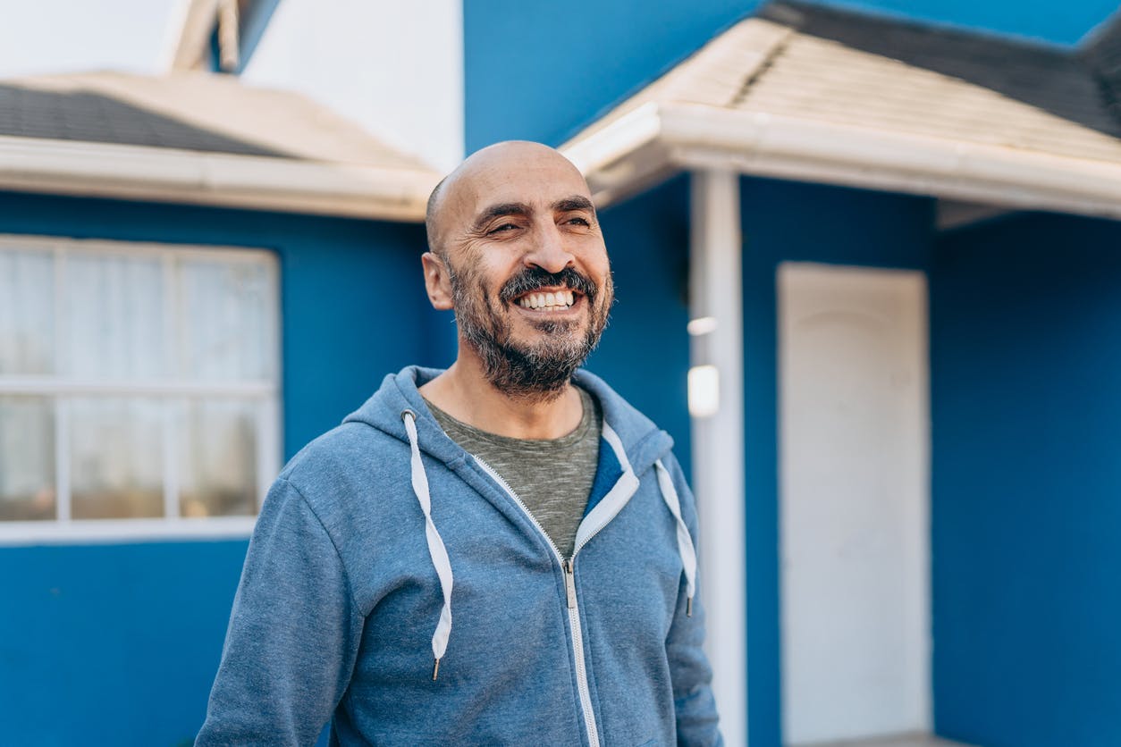 Man in blue zip up jacket smiling in front of a blue house